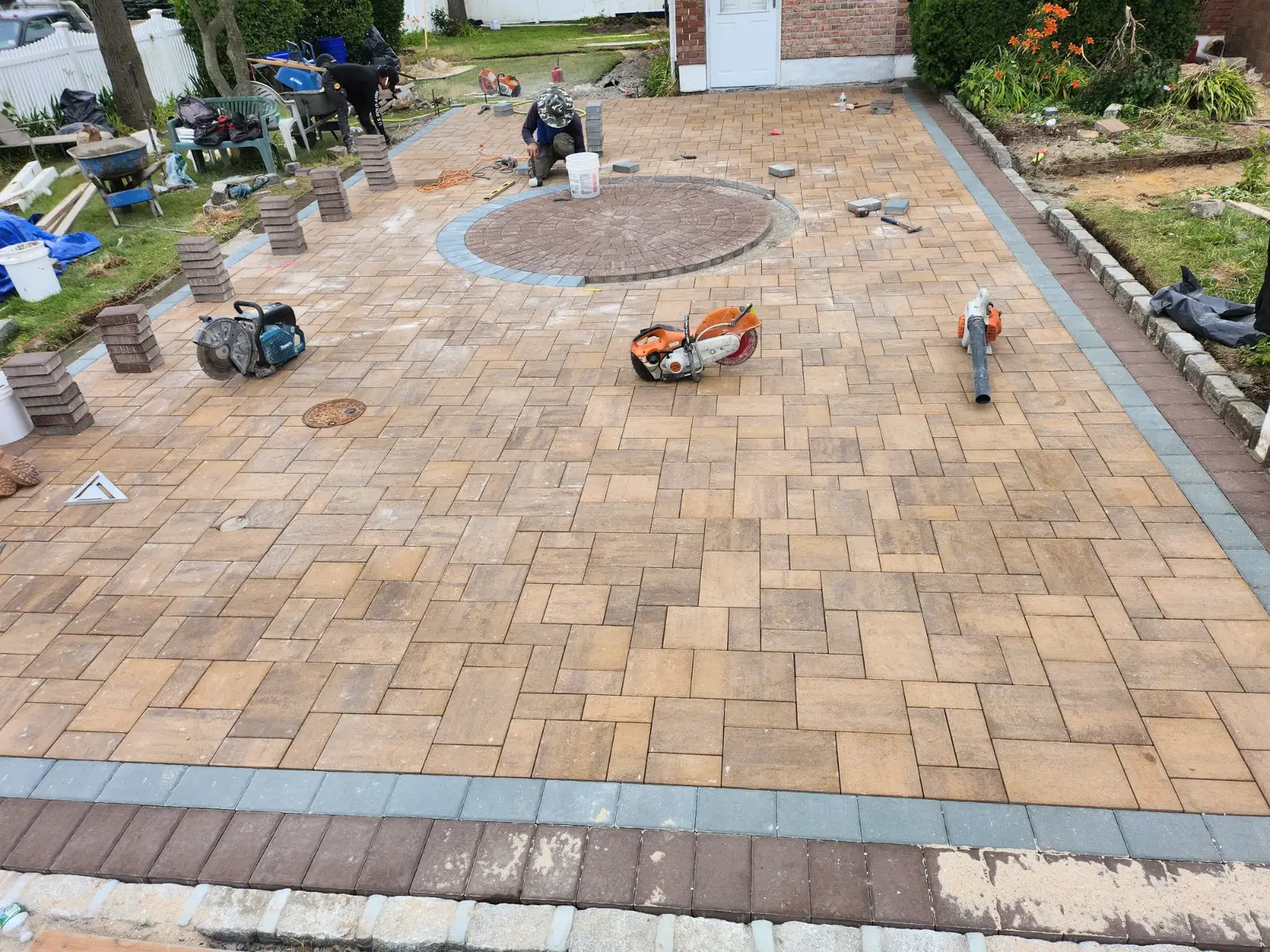 Promaster crew mid-installation of a paver patio with a circle inlay, tools and paver pallets on site, Long Island, NY